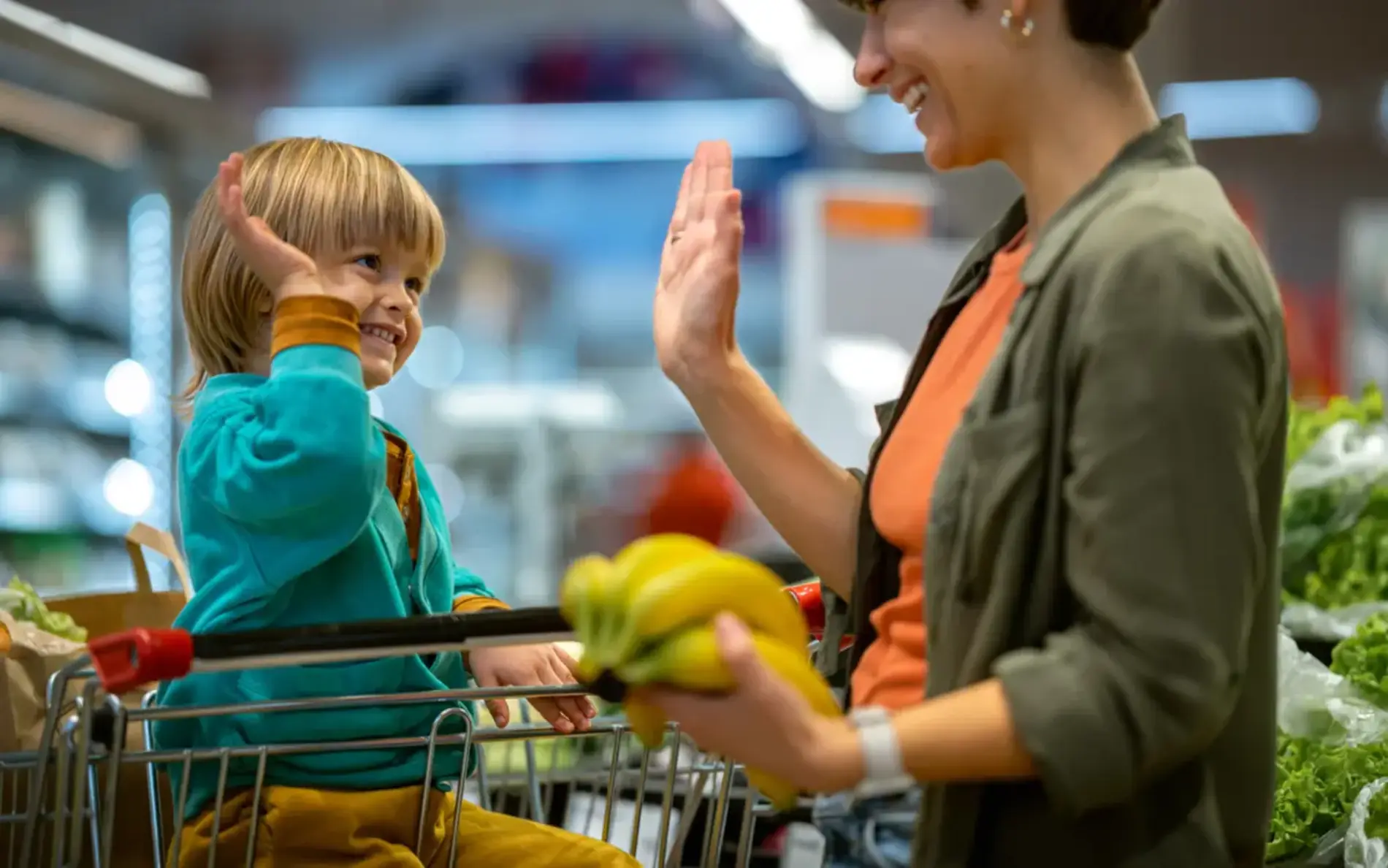 Kind im sitzt im Einkaufswagen und Mutter mit Paprika in der Hand steht daneben. Sie schauen sich in die Augen und schlagen High-Five