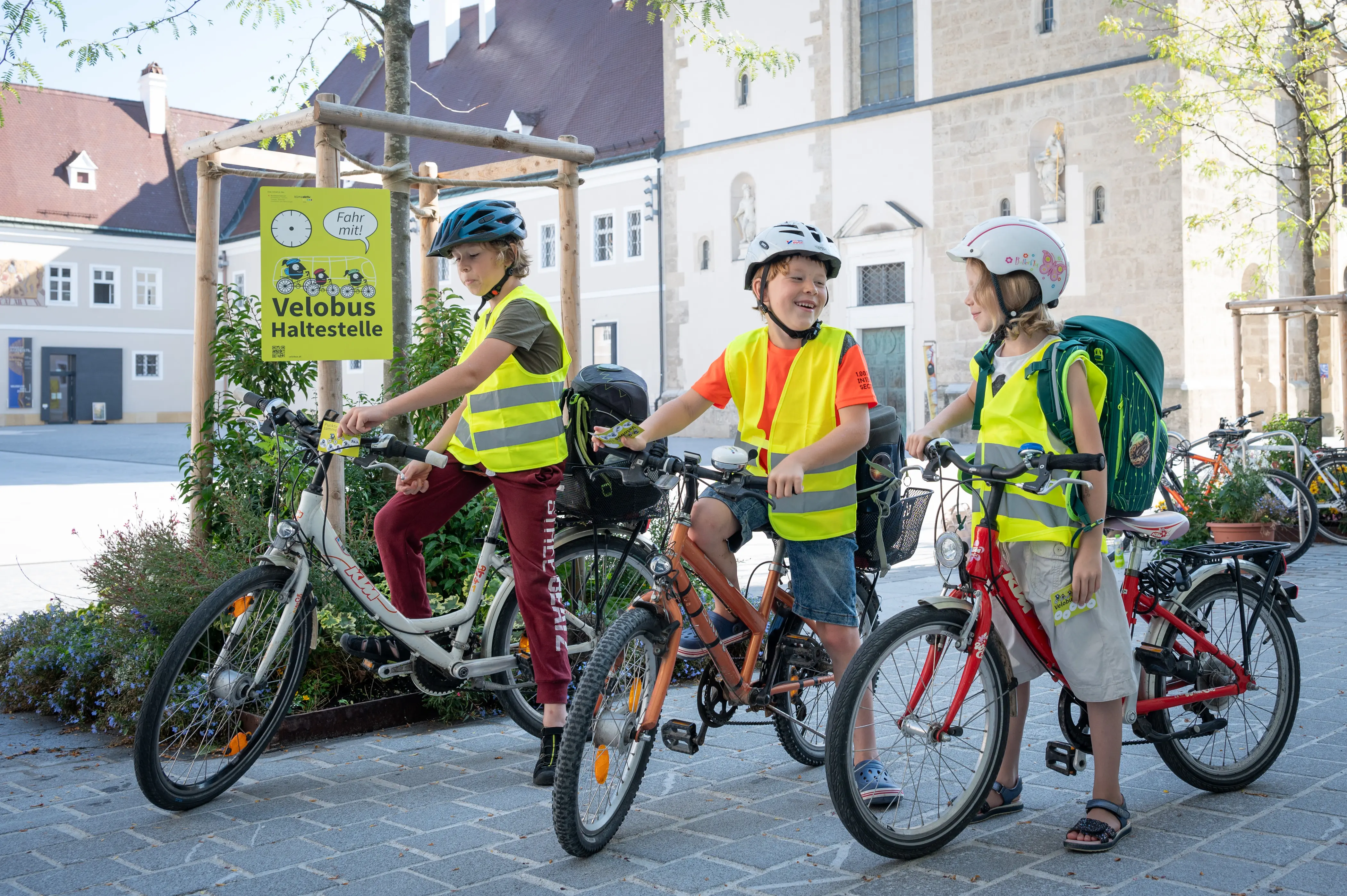 Drei Kinder auf dem Fahrrad mit leuchtender Schutzweste vor der Velobus Haltestelle