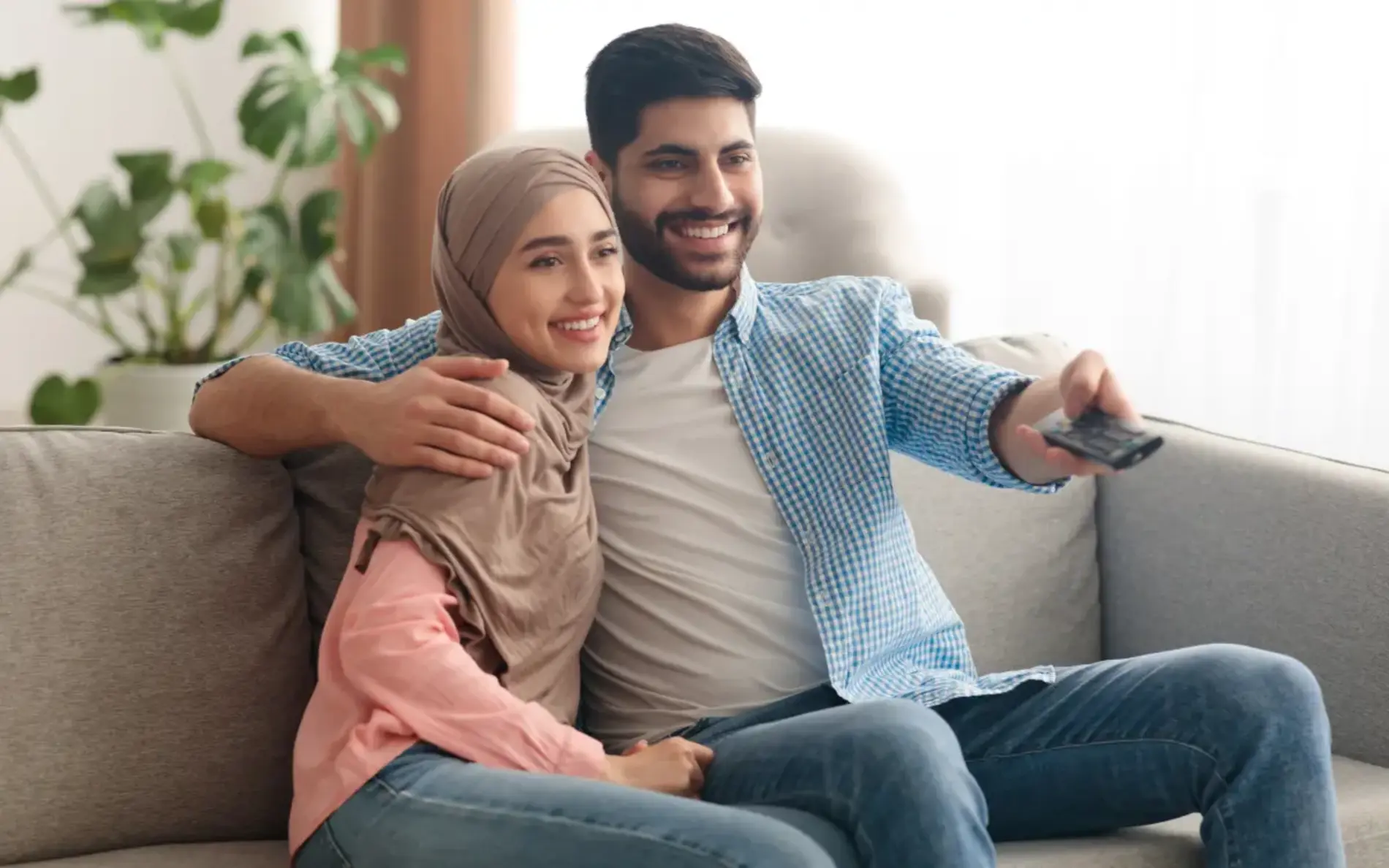 Eine Frau und ein Mann liegen Arm in Arm auf einer Couch. der Mann schaltet mit einer Fernbedienung den Fernseher an.