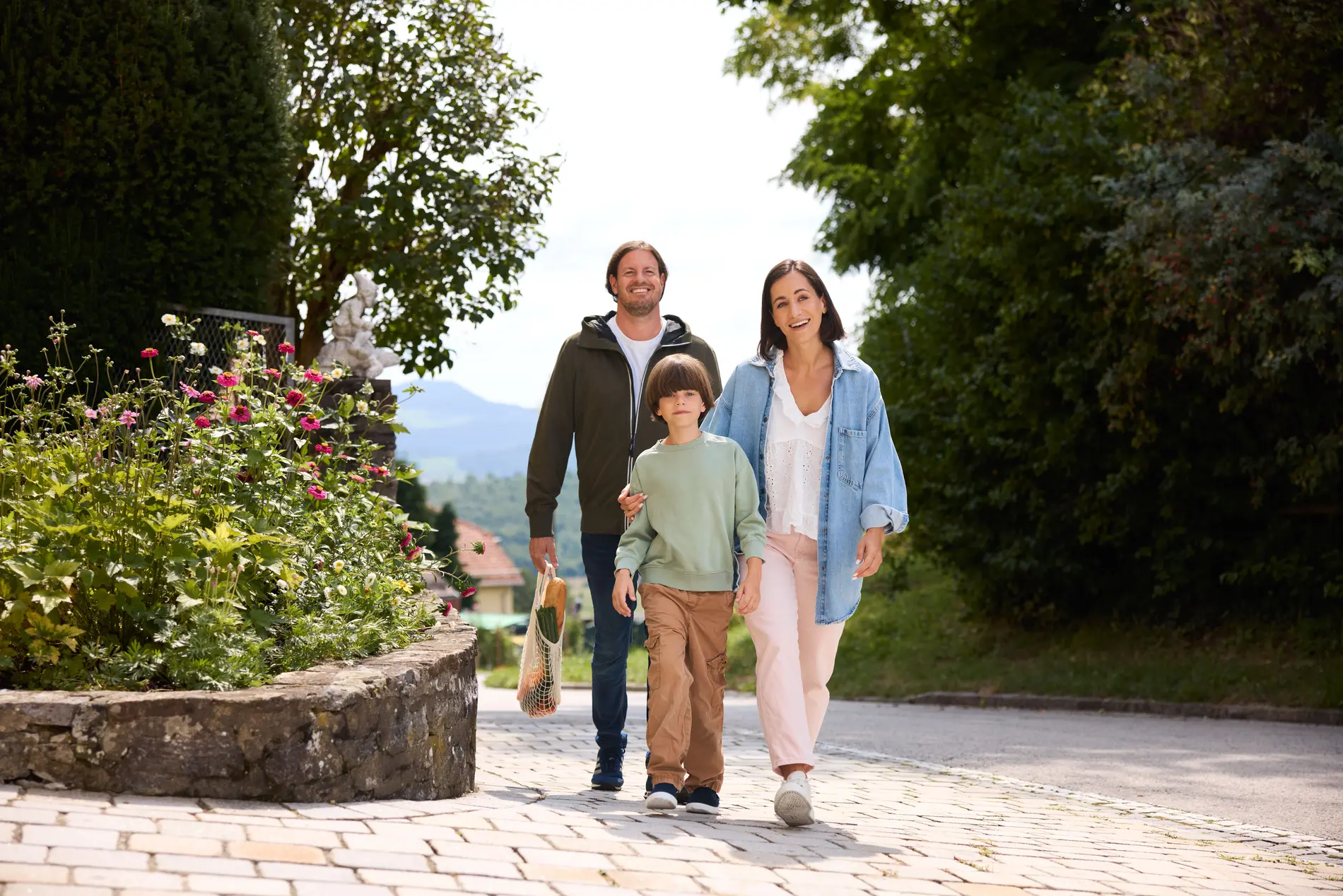 Eine Familie - ein Vater, eine Mutter und ein Volksschulkind - gehen auf einem gepflasterten Gehweg entlang. Links und rechts ist der Weg begrünt.