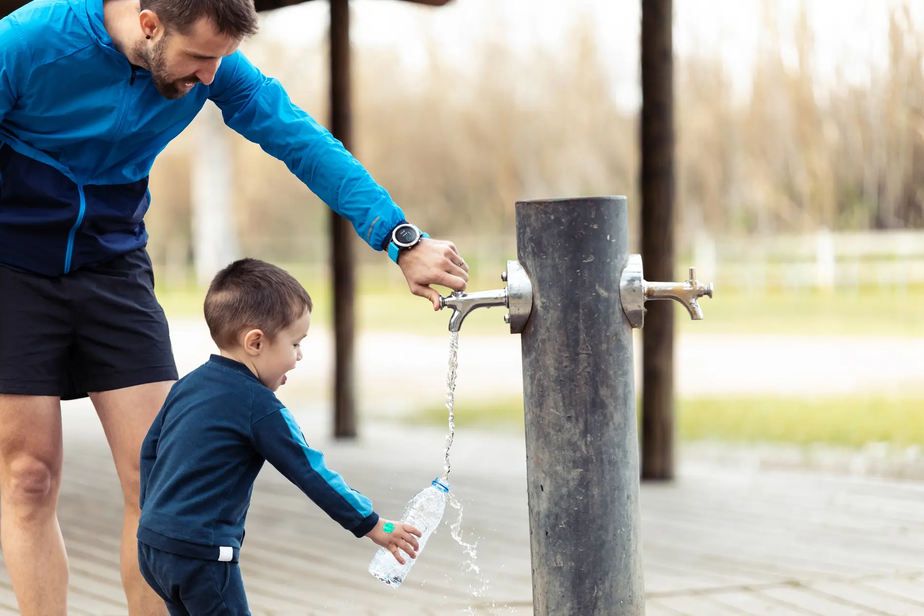 Vater öffnet Wasserhahn des Trinkbrunnes, während Kindergartenkind die Wasserflasche auffüllt.