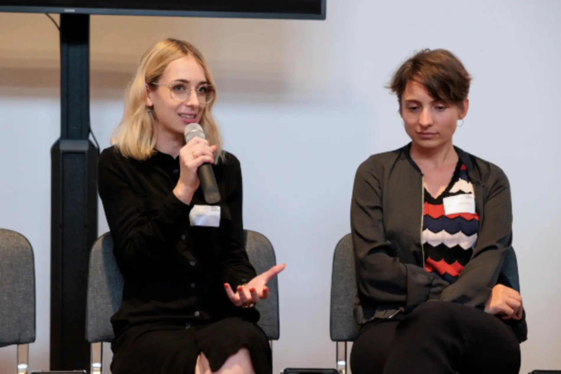 Verena Mischitz und Katharina Kropshofer sitzen am Podium. Mischitz spricht in ein Mikrofon zum Publikum.