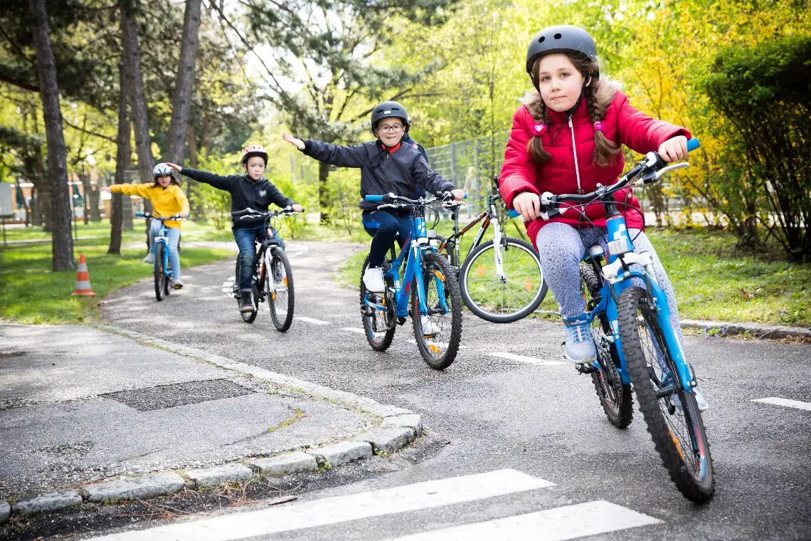 Vier Kinder fahren hintereinander auf der Straße mit dem Fahrrad und biegen in eine Straße ein. Der Radweg ist umgeben von einem Park. Die Kinder halten die rechte Hand ausgestreckt und signalisieren damit, dass sie rechts abbiegen.