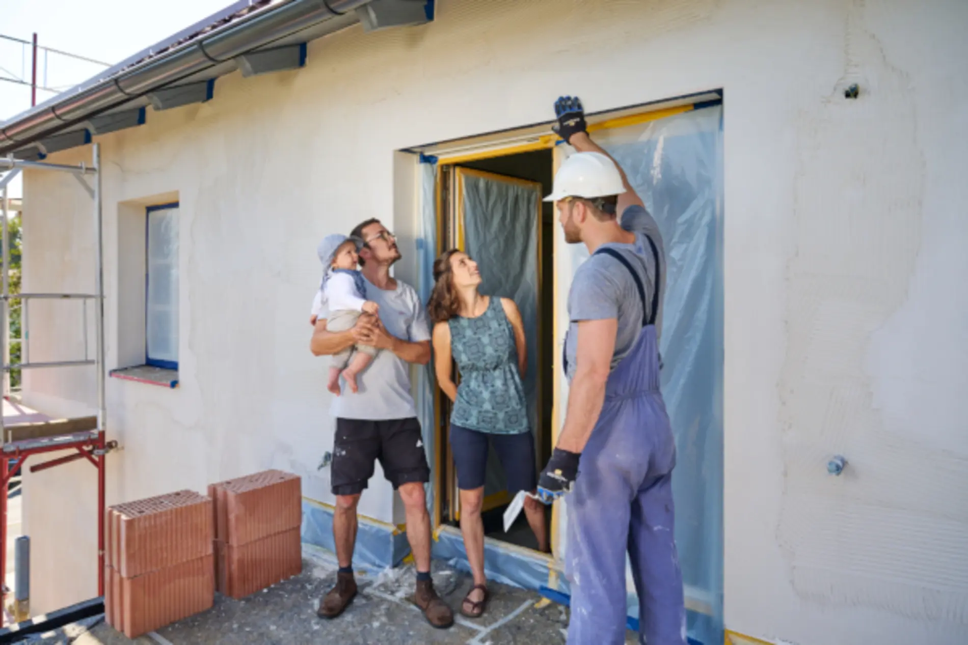 Vor einer tür mit Plastikfolie steht eine Frau, ein Mann mit Kind im Arm und ein Bauarbeiter. Der Bauarbeiter scheint etwas zu erklären, die drei anderen Personen schauen dabei zum Türstock.