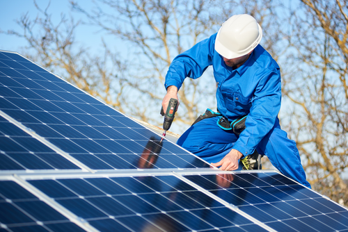 Ein Mann mit Helm und blauen Overall befestigt mit einem Akkuschrauber gerade die PV-Anlage