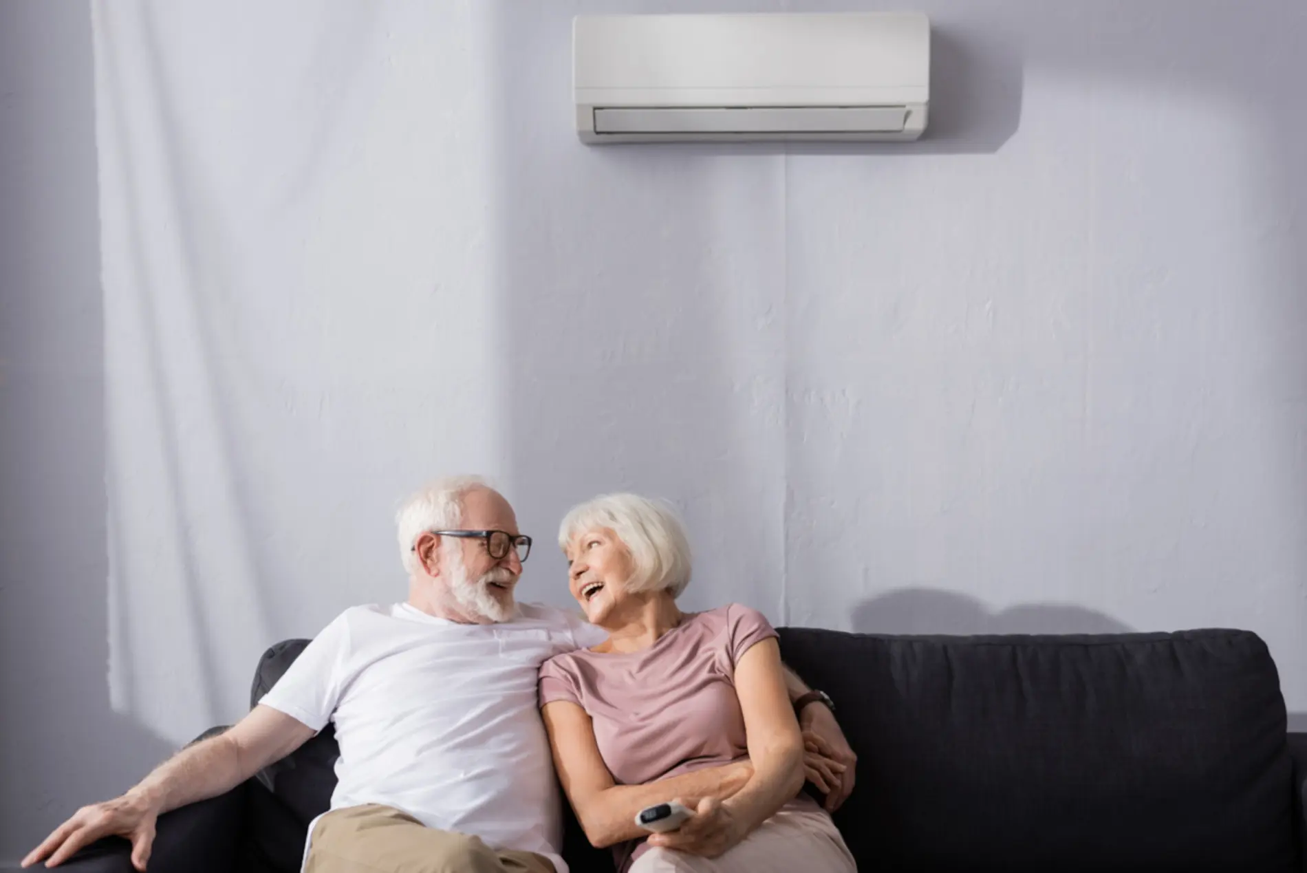 Ein älteres Paar sitzt auf einem Sofa uunter einer Klimaanlage. Der Mann nimmt dabei seine Frau in den Arm während diese ihn anlächelt. 