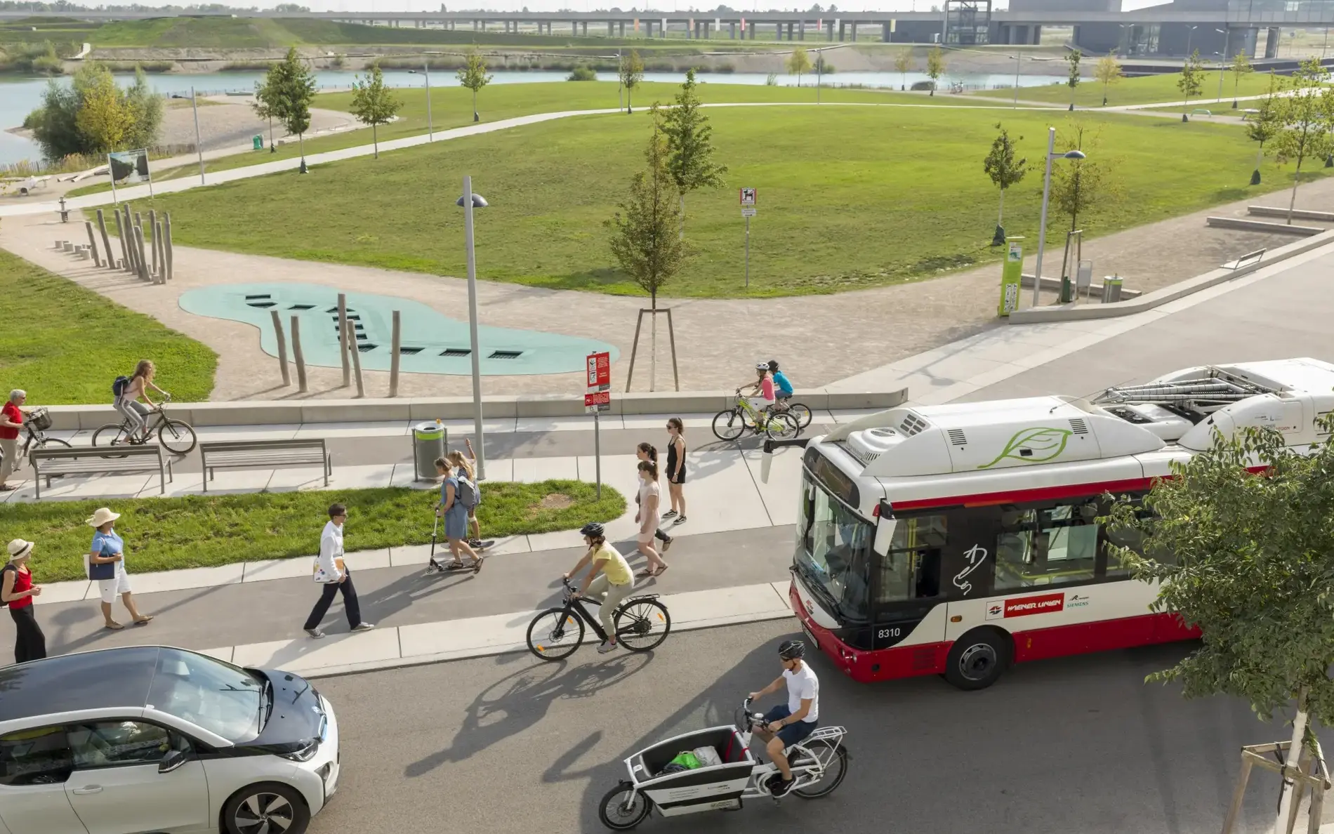 Ansicht von oben über eine Straße mit anliegendem Gehweg und Radweg. Zu sehen sind Räder, ein Transportrad, Bus, Elektroauto, viele zu Fuß gehende