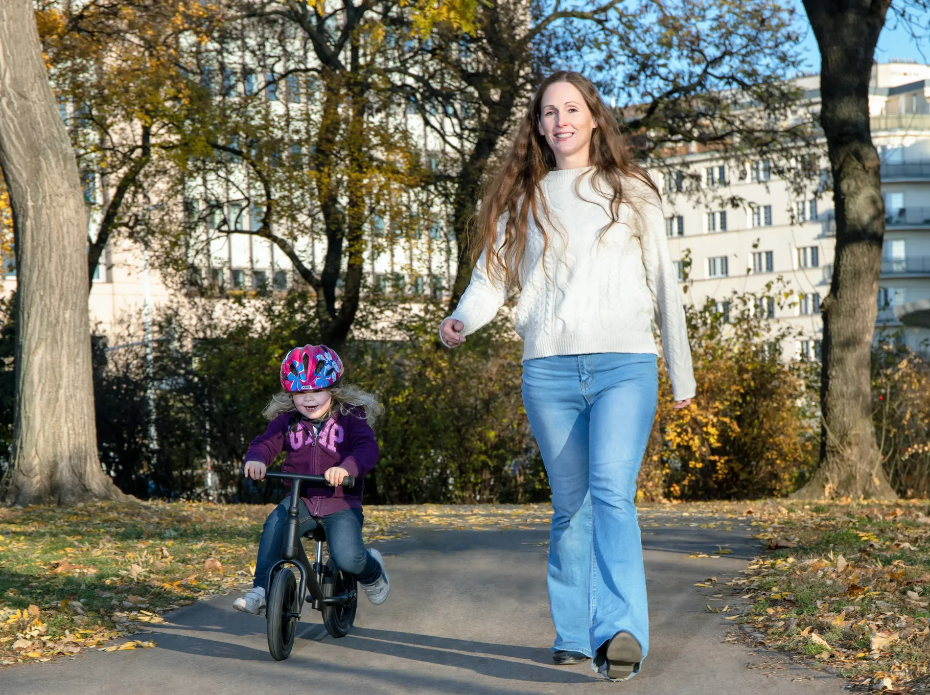 Kind auf Laufrad und Mutter gehend daneben durch einen Park in städtischer Umgebung