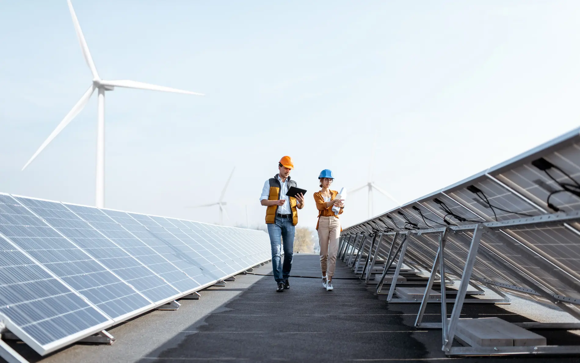 Zwei Personen mit Helm gehen durch zwei lange PV-Anlagen durch, im Hintergrund Windräder