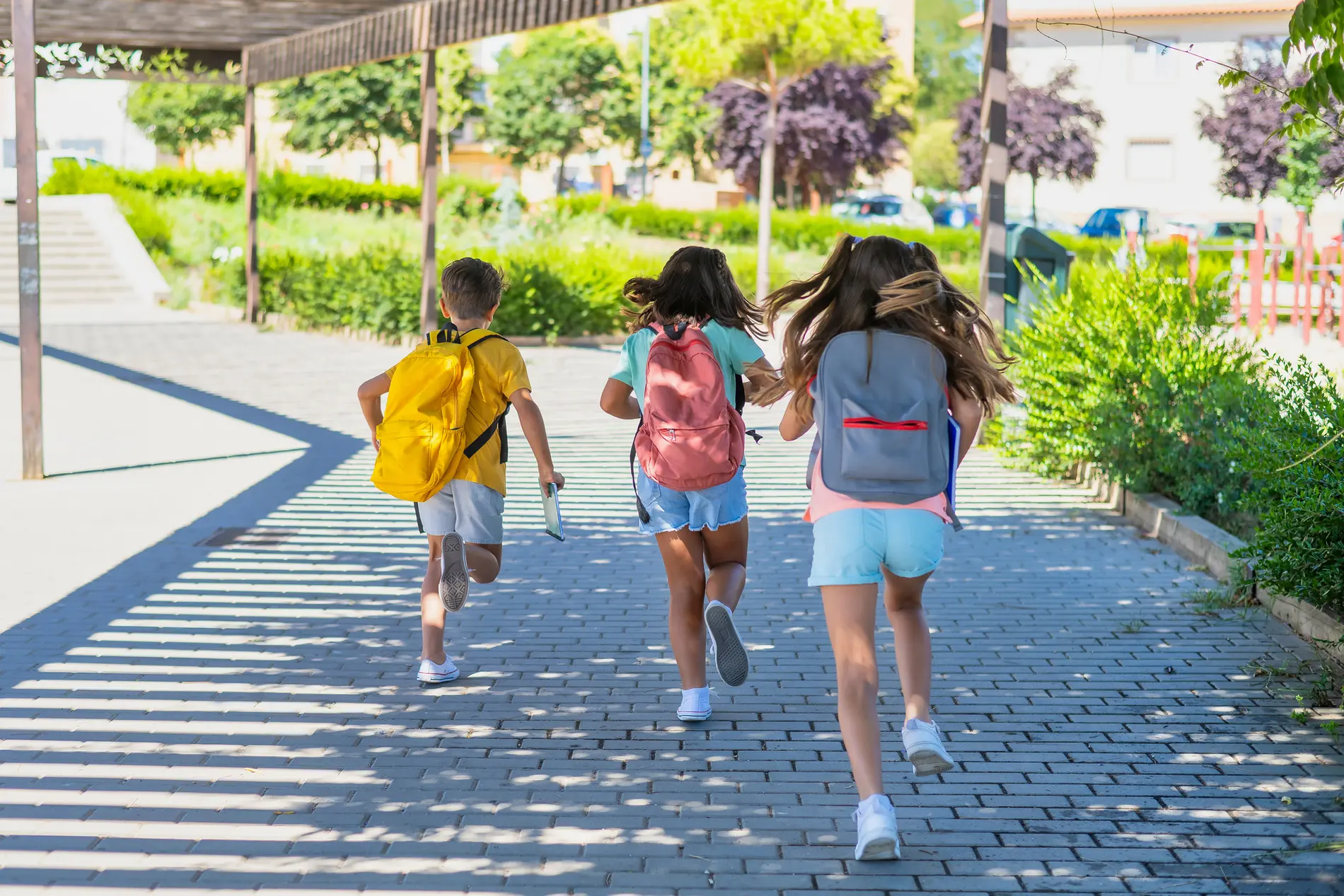 Kinder laufen mit Schultaschen am Rücken entlang eines breiten Gehweges, beschattet von einer Pergola. Rechts davon Begrünung.