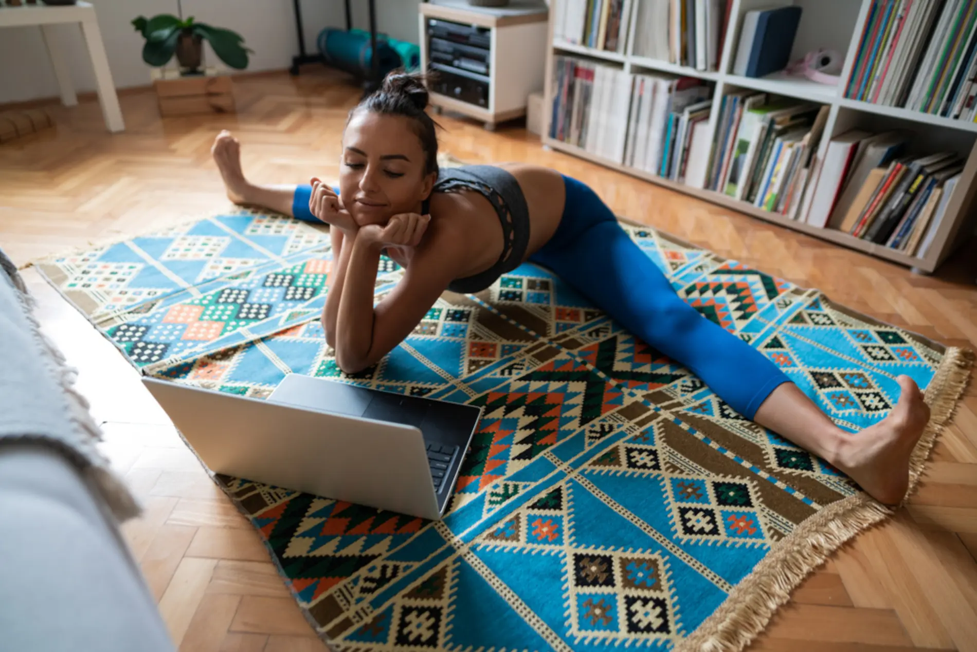 Zu sehen ist eine Frau, die auf einem Teppich vor ihrem Laptop einen Spagat macht. 