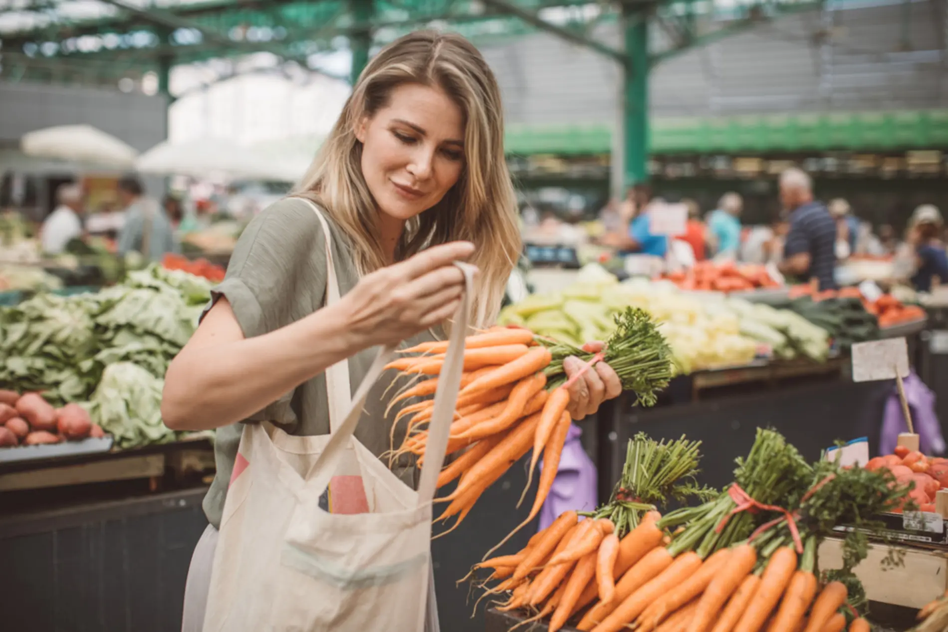 Frau mit Stoffsackerl am Gemüsemarkt. Sie packt einen Bund Karotten ein.