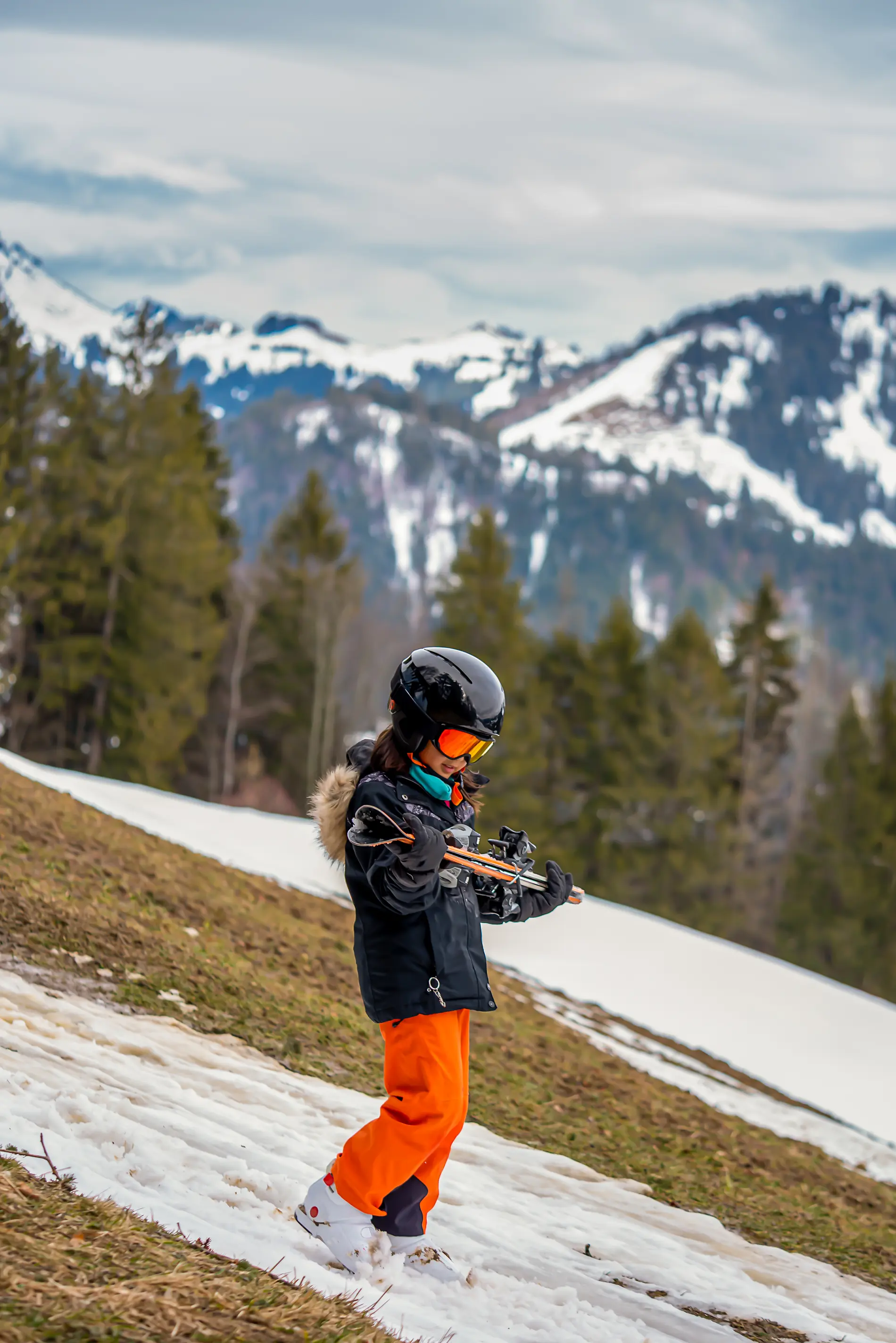 Ein Kind geht im Skianzug mit den Skiern in den Händen an einem kleinen weißen Schneeband hinunter. Im Hintergrund sieht man, dass die Skipiste in weiten Teilen grün ist.