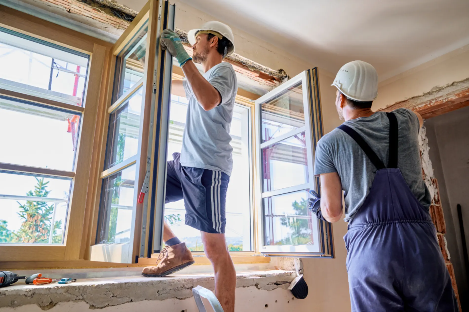 Bei einem offenen Fenster steht ein Bauarbeiter und baut ein Fenster ein. Ein anderer Bauarbeiter ist ihm behilflich. 