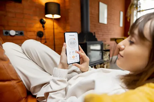 Eine Frau mit braunen Haaren sitzt auf der Couch und steuert mittels einer Handyapp ihre Smarthomeanwendungen